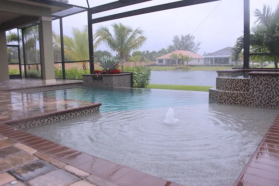 An infinity-edge pool with a bubbler fountain overlooks a lake under a screened lanai in the rain.