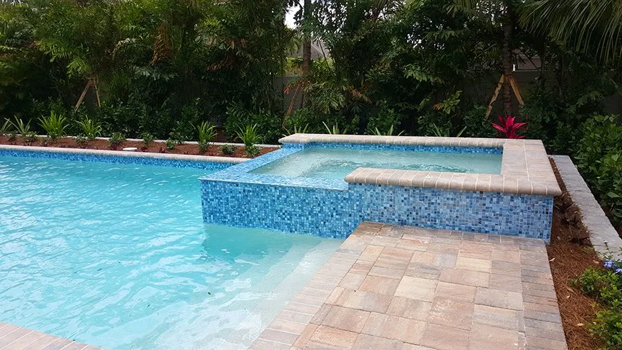 Rectangular pool with a raised glass-tile spa spilling into the pool, set on a paver patio surrounded by lush landscaping.