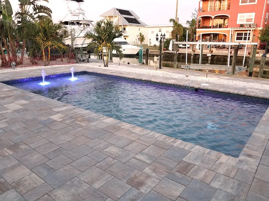 A rectangular backyard pool with two lit water bubblers at dusk on a canal lined with boats.