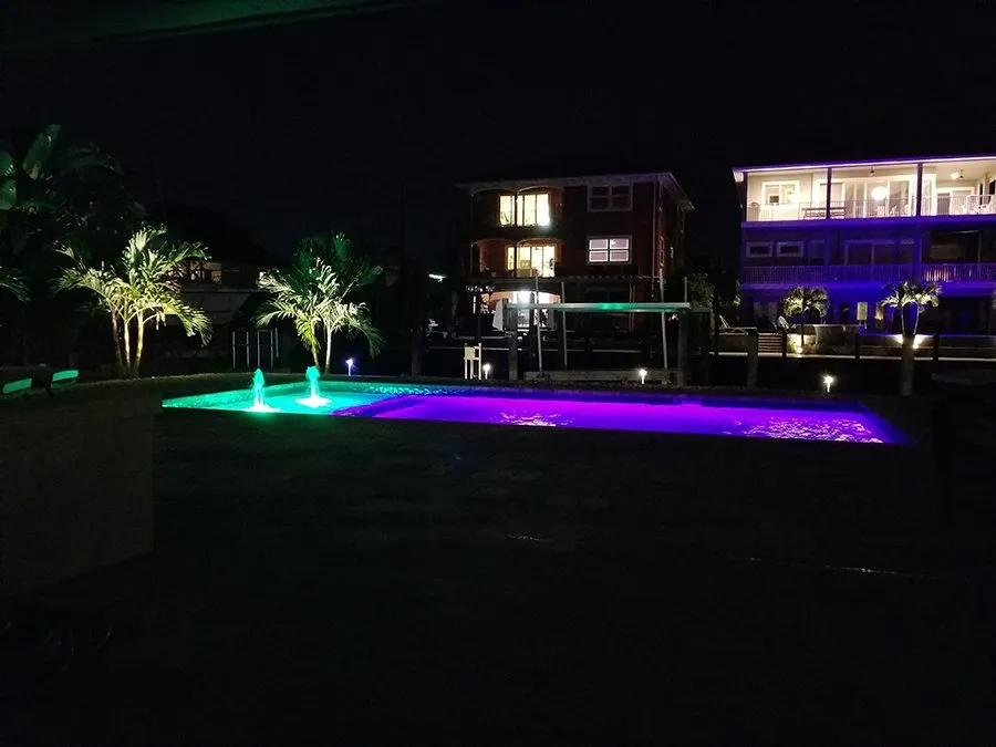 Nighttime rectangular pool lit with vibrant green and purple LED lighting and bubbler jets, waterfront homes in background.