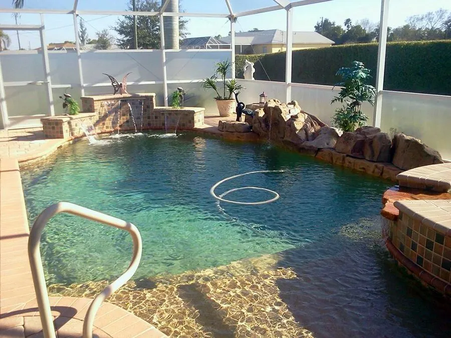 A freeform backyard pool with a rock waterfall feature and raised spa under a screened enclosure.