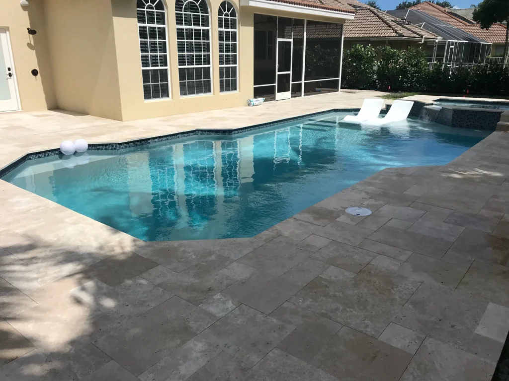 A geometric pool with raised spa and in-water loungers on a travertine paver patio behind a stucco home.