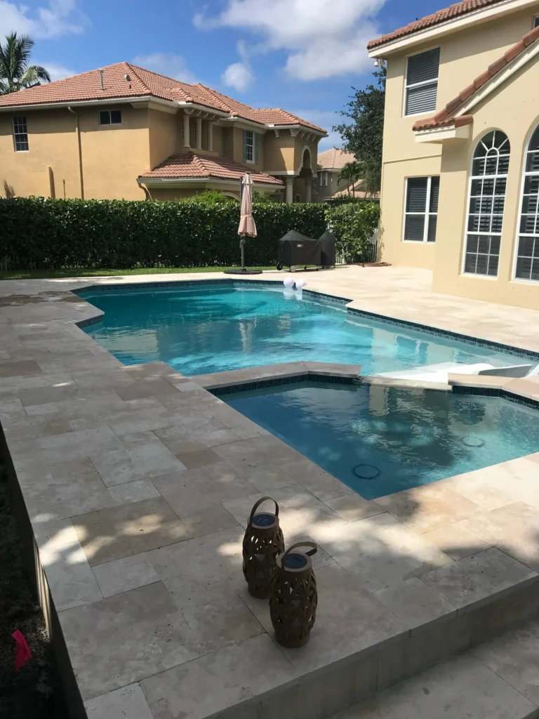 An L-shaped backyard pool with an integrated spa and travertine deck between two large homes.