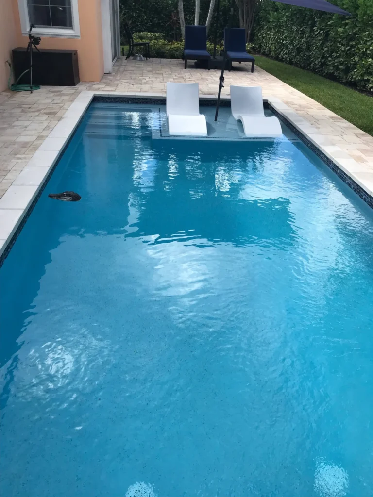 A small rectangular backyard pool with two white in-pool loungers on a sun shelf and travertine deck.