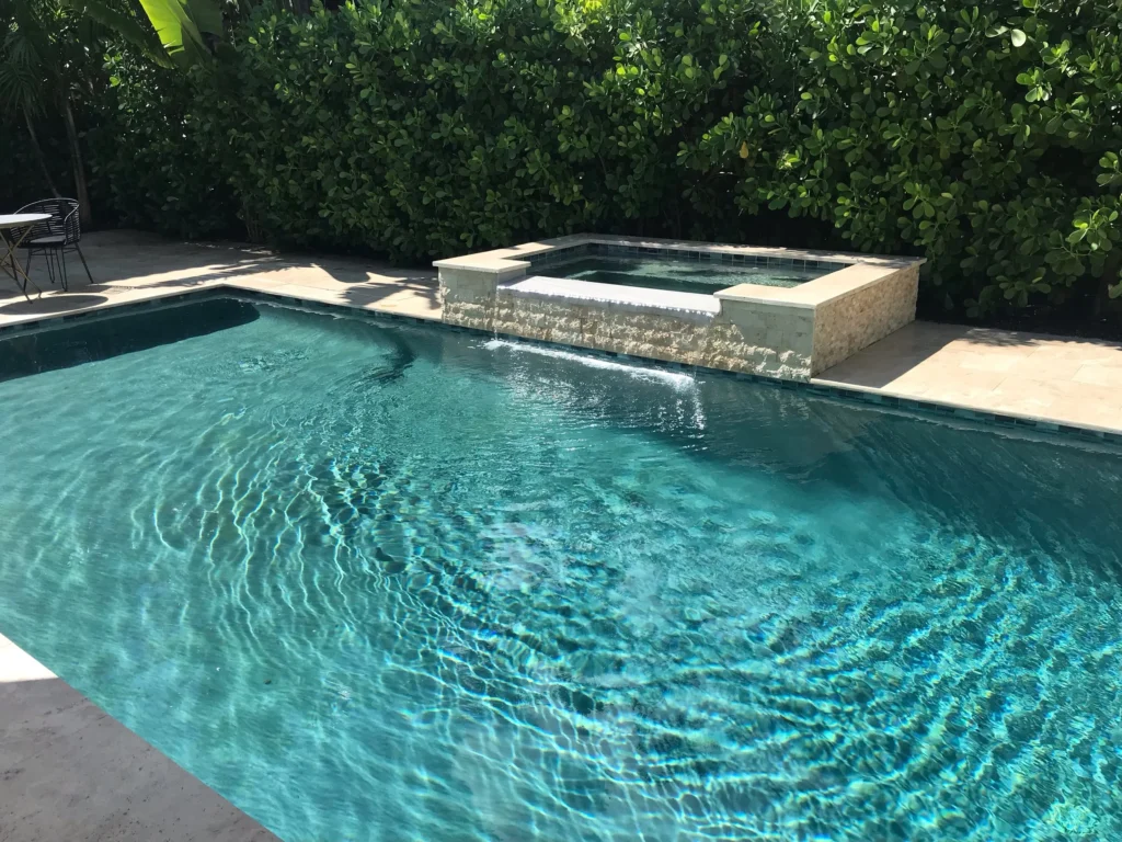 A rectangular backyard pool with a raised stone spa spilling water into it, surrounded by lush hedges.