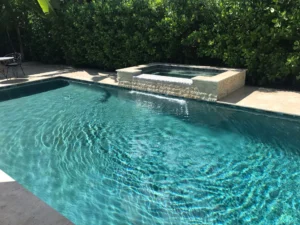 A rectangular backyard pool with a raised stone spa spilling water into it, surrounded by lush hedges.