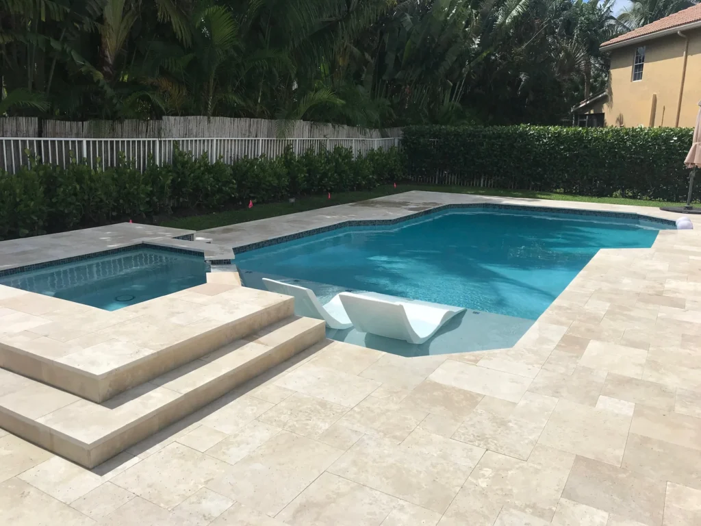 A geometric backyard pool with an attached raised spa and two in-pool loungers on a travertine paver patio.