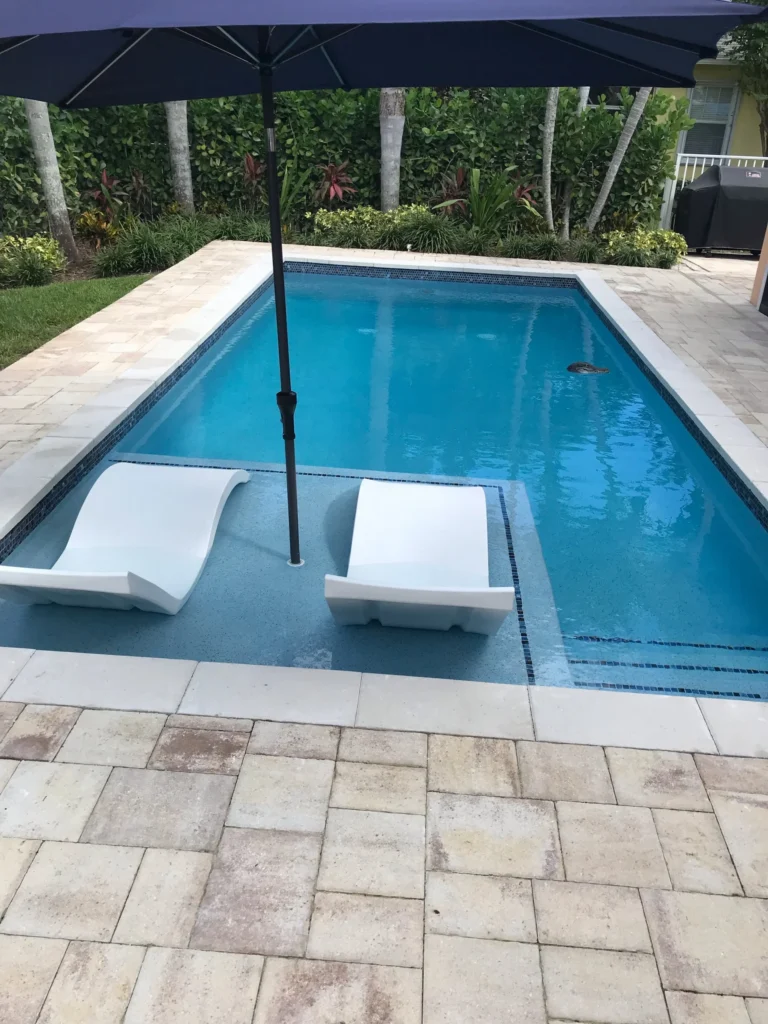 Rectangular pool with a sun shelf holding two white in-pool loungers under a large umbrella.