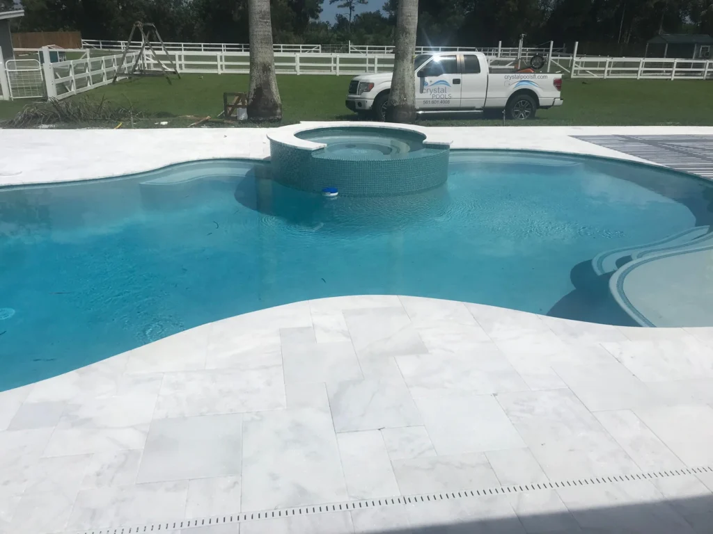 Freeform pool with a raised tiled spa and travertine paver deck, white fencing and palm trees in the background.