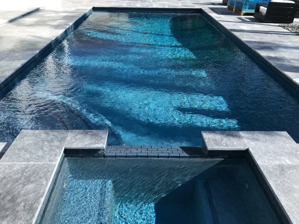 A dark-bottomed rectangular pool with a raised spillover spa and stone paver deck.