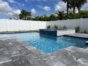A rectangular backyard pool with a raised spa clad in mosaic tile and deck jets arcing water, surrounded by gray travertine pavers and a white vinyl fence.