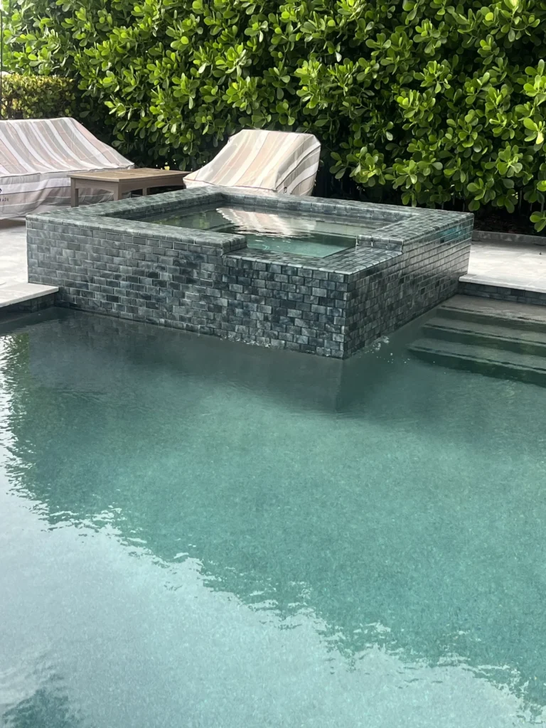 A raised tile-clad spa sits at the edge of a pool with loungers and greenery behind.