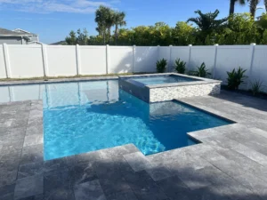 A geometric pool with a raised tiled spa spilling into it, surrounded by gray travertine decking and a white vinyl fence.