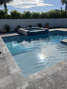 A rectangular backyard pool with a raised tiled spa spilling into the pool, surrounded by a travertine deck and white vinyl fence.