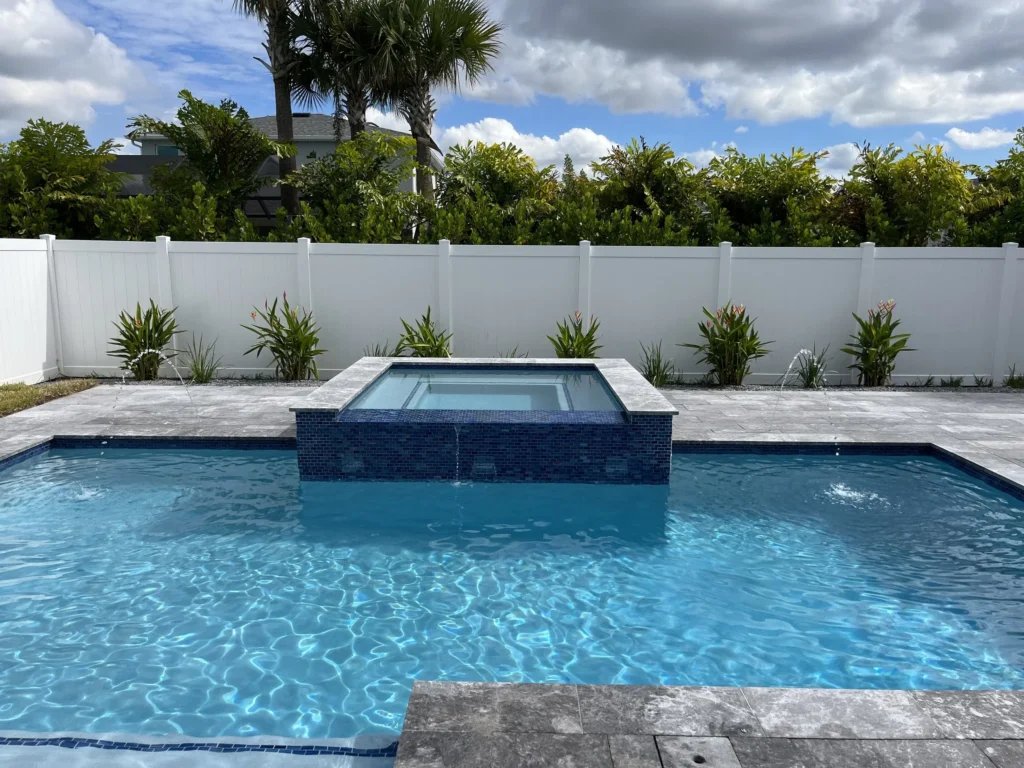 A rectangular backyard pool with a raised tiled spa spilling over into the pool and deck jets arcing water, enclosed by a white vinyl fence.