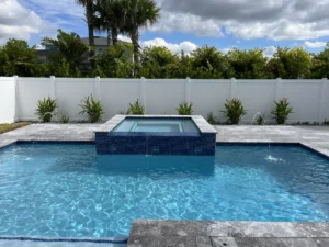 A rectangular backyard pool with a raised tiled spa spilling over into the pool and deck jets arcing water, enclosed by a white vinyl fence.