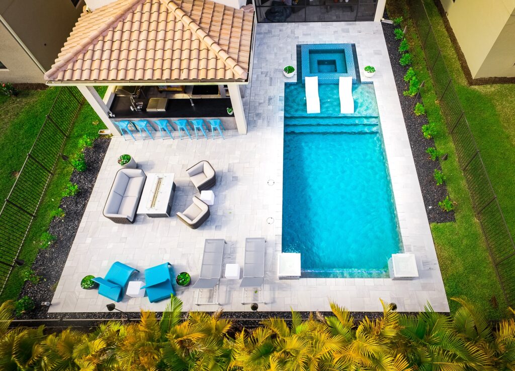Aerial view of a rectangular backyard pool with a raised spa, in-water loungers, and a stone patio with an outdoor kitchen pavilion.