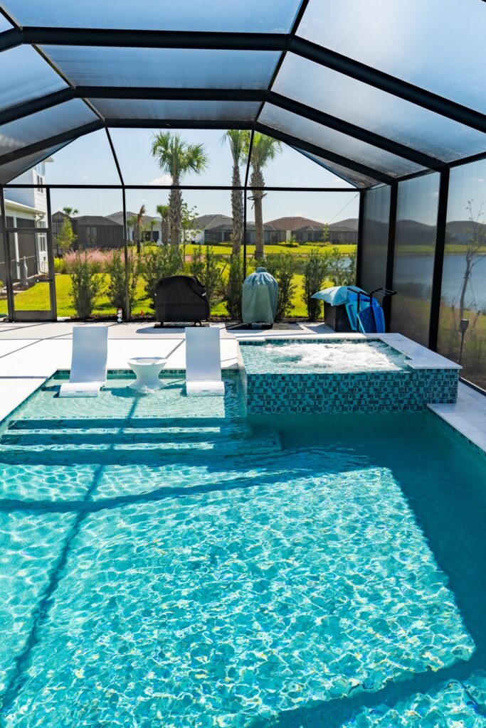 A screened pool with a tanning ledge, in-pool loungers, and a raised spa overlooking a lake with palm trees.