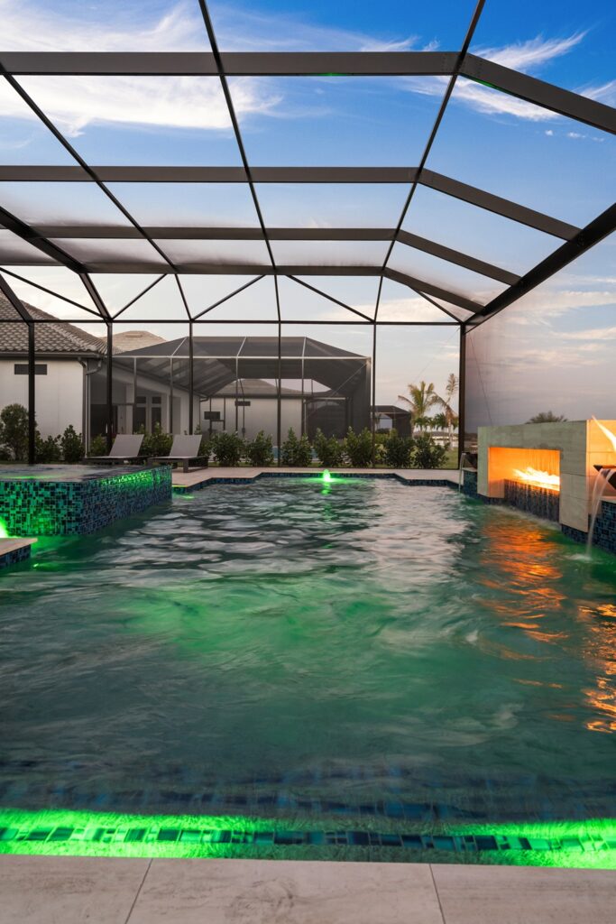 A screened pool at dusk lit with green LED lighting and a stone fireplace with dancing flames beside a water scupper.