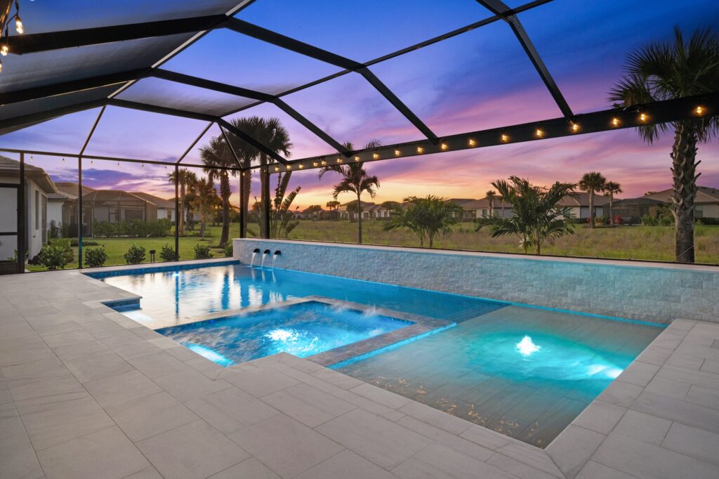 A multi-level pool and raised spa glow with blue LED lighting under a screened enclosure at dusk, with a stone accent wall and open field view beyond.