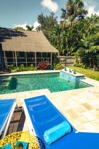 A small rectangular backyard pool with a raised spa featuring three water bubblers, surrounded by travertine pavers and tropical landscaping.