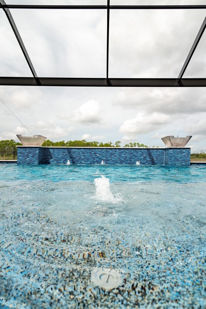 A raised tiled water wall with two scupper bowls feeds a pool under a screened enclosure on an overcast day.