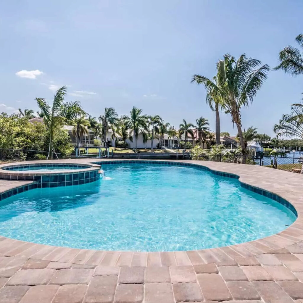 Freeform pool with attached raised spa and paver deck overlooking a canal lined with palm trees.