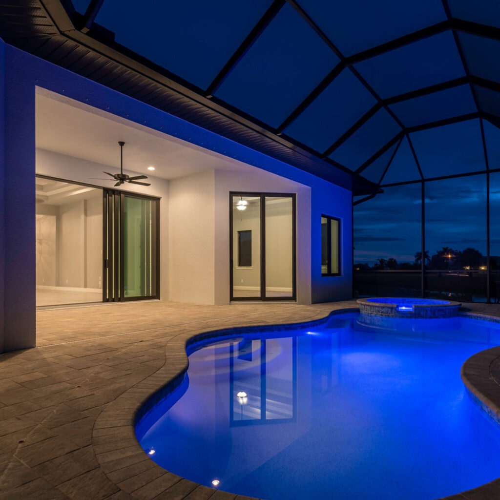 A freeform pool with blue LED lighting and a raised spa under a screened enclosure at night.