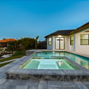A raised tiled spa glows at dusk with a rectangular pool and canal-front backyard behind it.