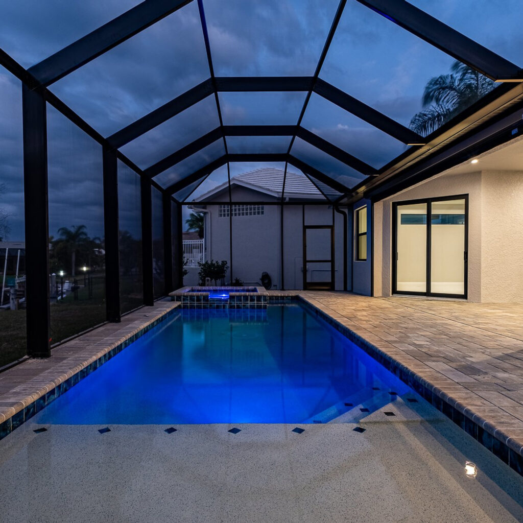 A screened rectangular pool with a raised spa glows blue at dusk beside a paver patio.