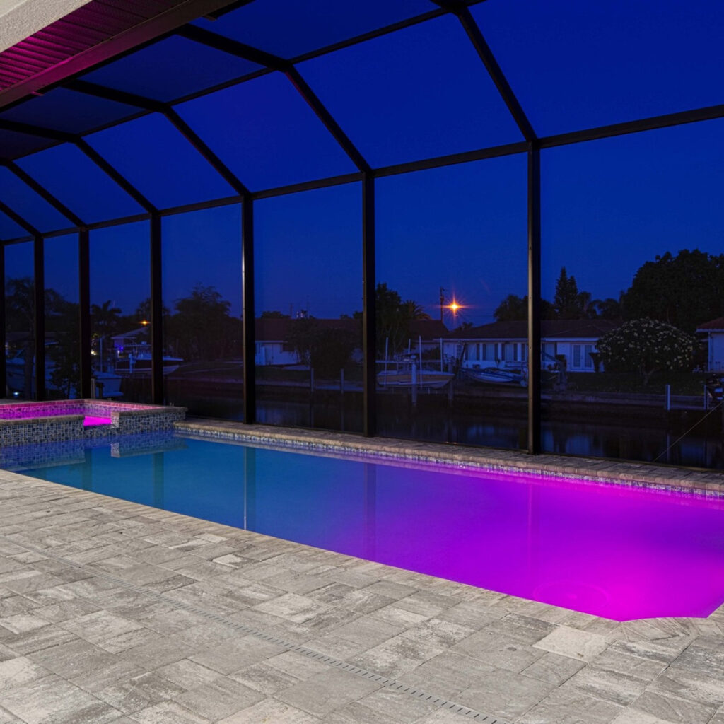 A screened backyard pool glowing with purple LED lighting at night beside a canal with docked boats.