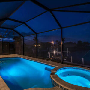 A screened pool with a round raised spa glowing blue at night beside a canal.