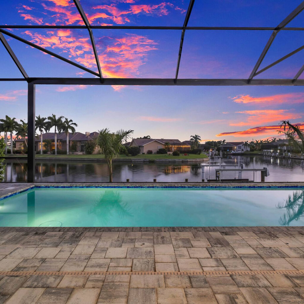 A screened pool overlooks a canal at sunset with vivid pink and orange clouds.