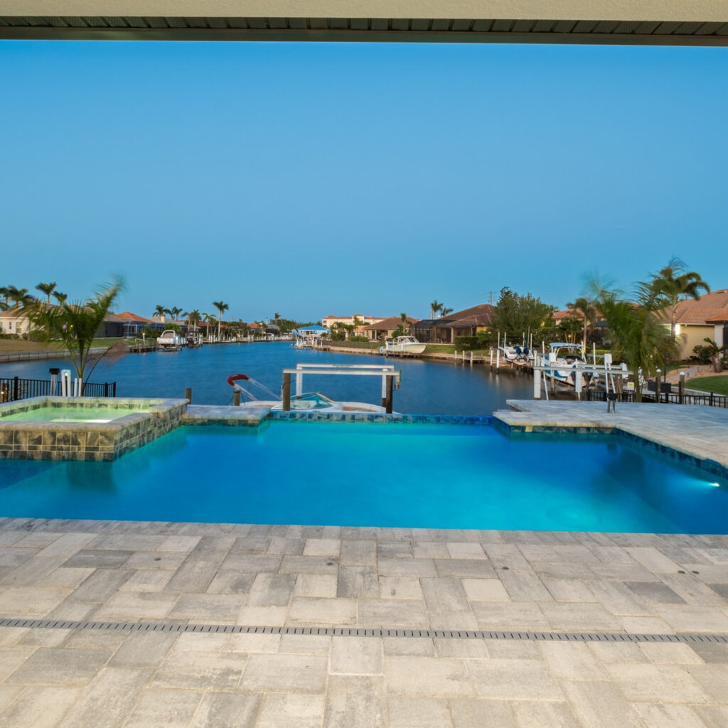An infinity-edge pool and raised spa overlook a canal lined with docked boats at dusk.