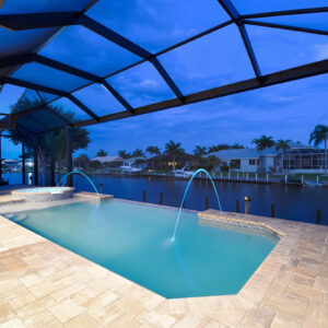 Rectangular pool with raised spa and twin deck jets under a screened enclosure overlooking a canal at dusk.