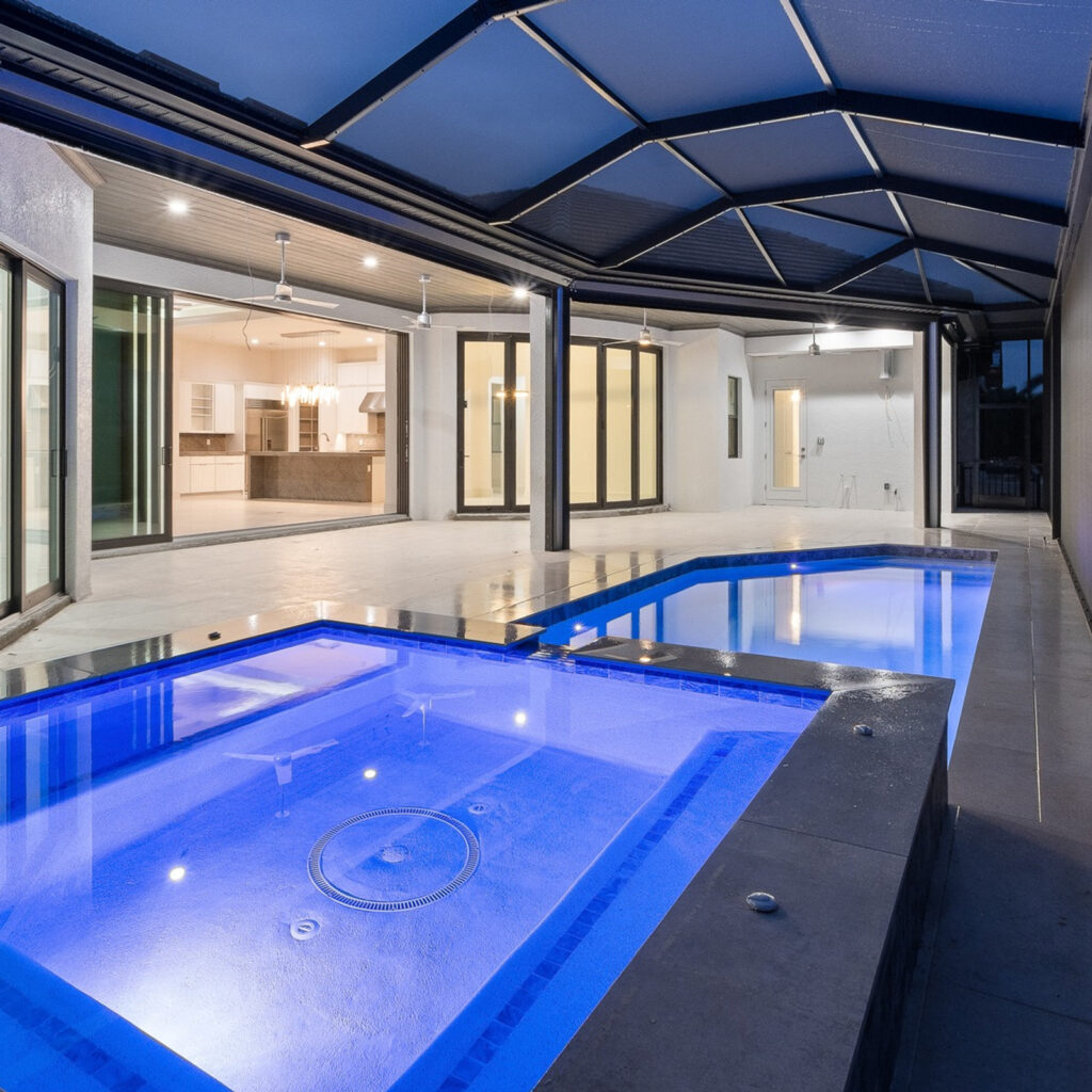 A raised spa glows blue at night in the foreground with a screened pool and modern home visible behind it.