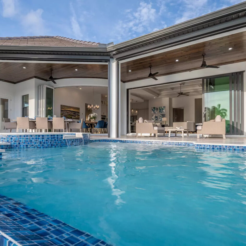 Blue mosaic-tiled pool edge in front of a luxury home with retractable glass walls opening to the living area.