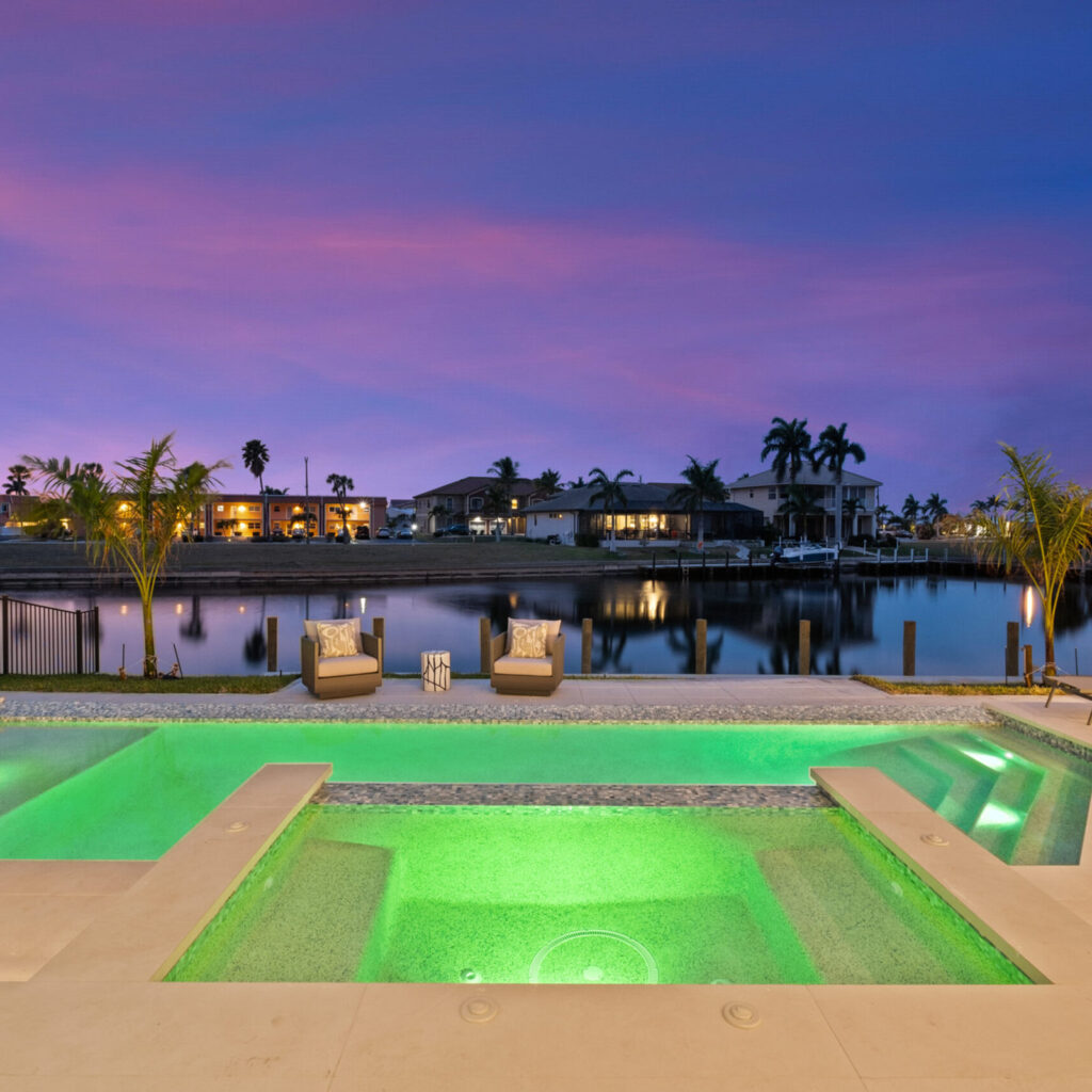 A raised spa glowing green at dusk overlooks an infinity-edge pool and a calm canal lined with palm trees and homes.