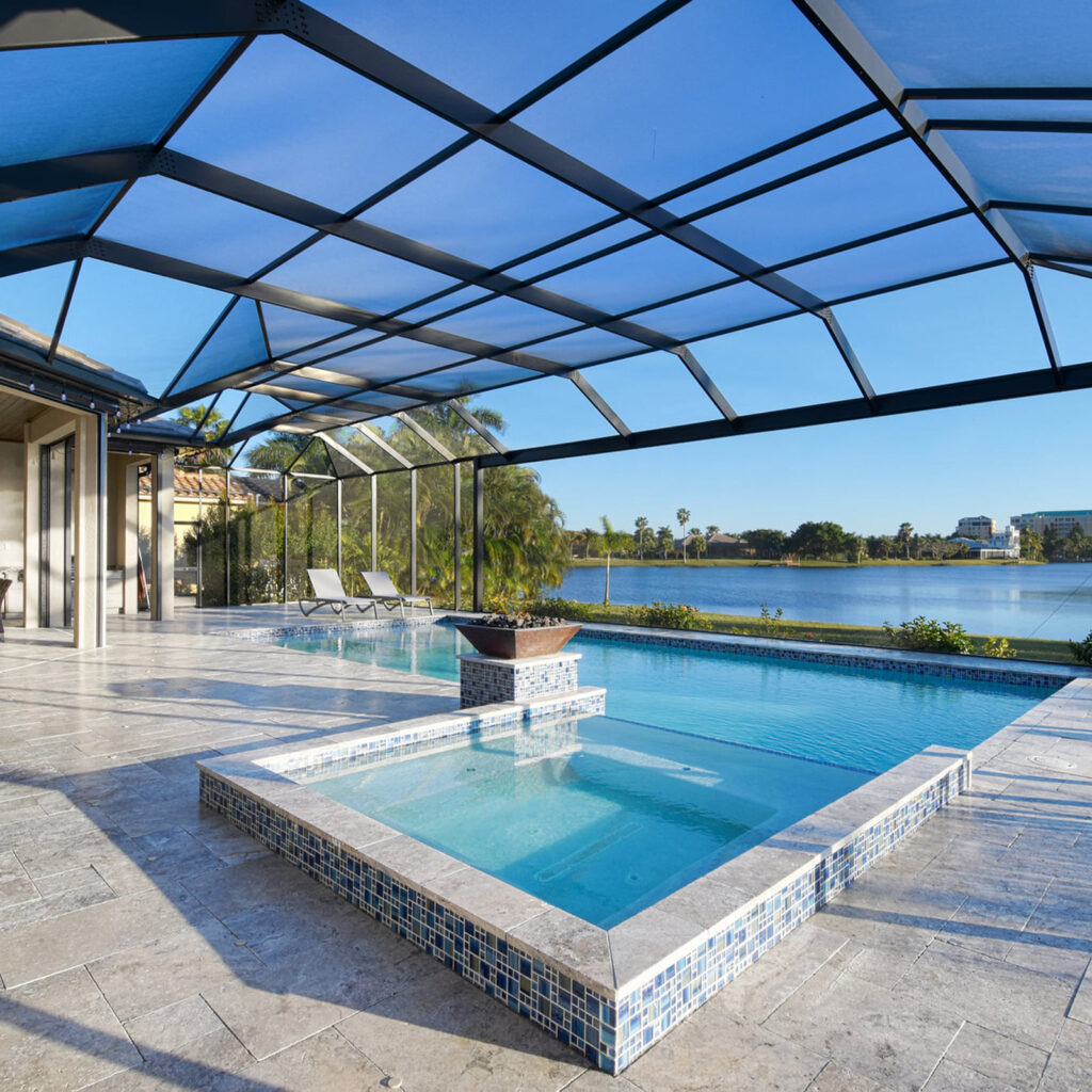 Screened lakefront pool with raised tiled spa and lit fire bowl on a travertine patio.