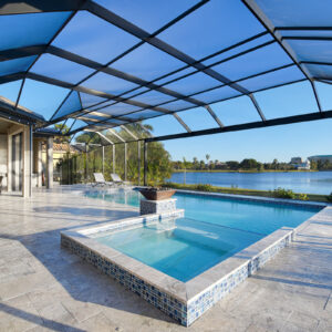 Screened lakefront pool with raised tiled spa and lit fire bowl on a travertine patio.