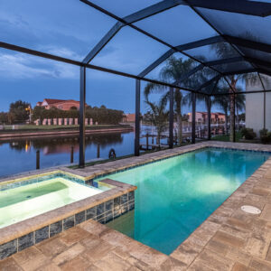 A screened rectangular pool with a raised spa overlooks a calm canal at dusk with palm trees and a dock.