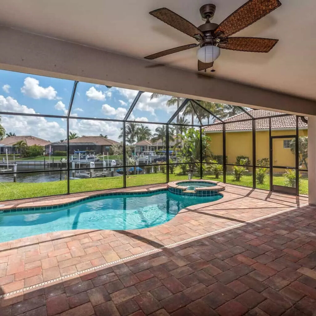 A screened lanai with a freeform pool and raised spa overlooking a canal lined with palm trees.