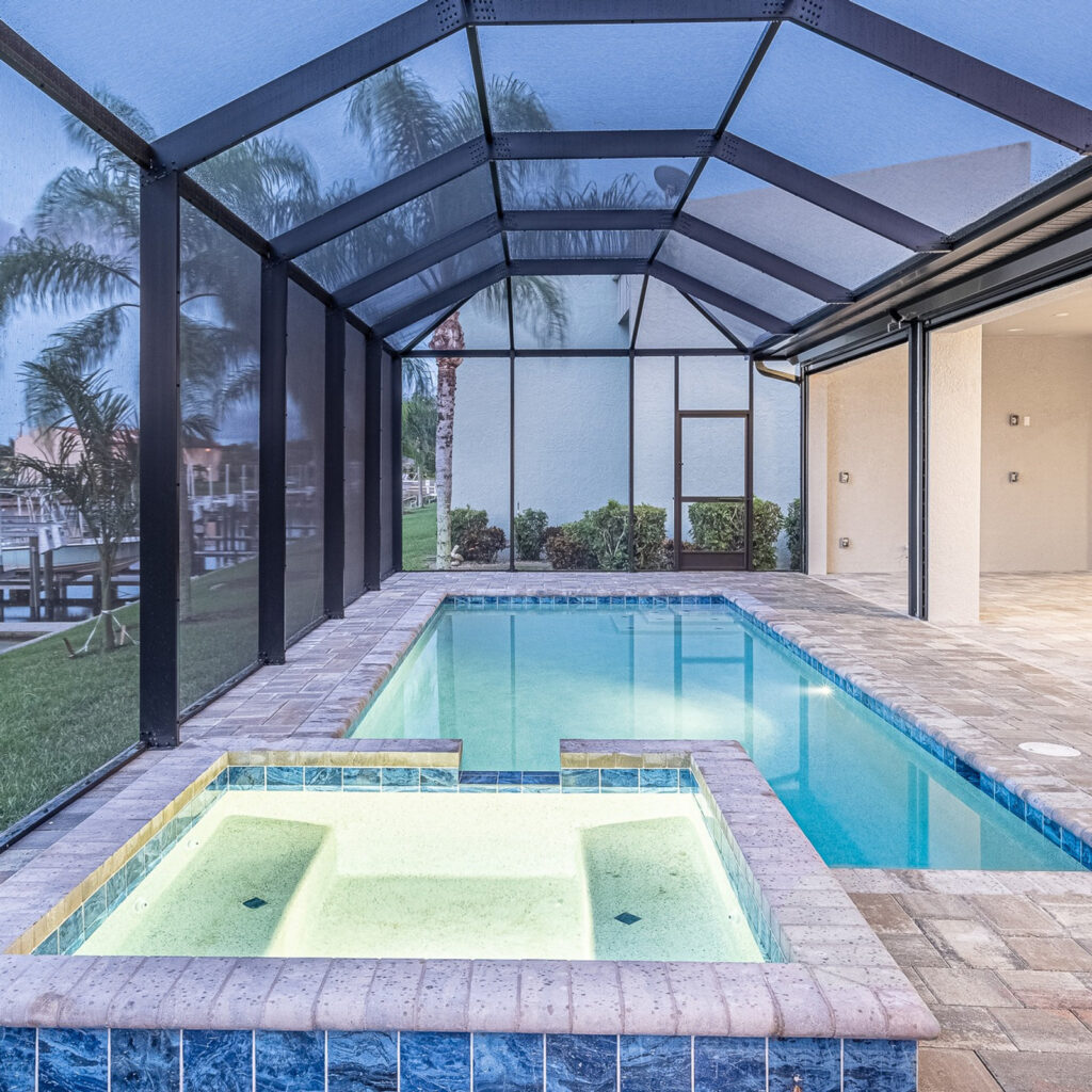 A raised tiled spa in the foreground of a screened pool enclosure at dusk with a canal visible beyond.