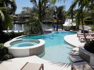 A freeform pool with a raised spa and waterfront canal view, surrounded by loungers and palm trees.