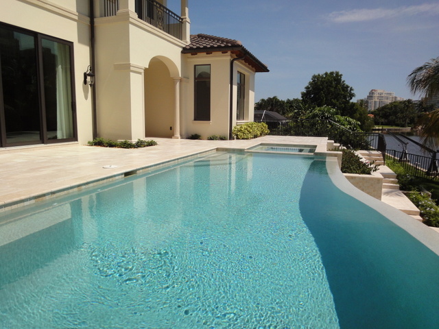 A curved infinity-edge style pool with a raised spa beside a luxury home overlooking a canal.