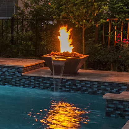 A raised fire bowl on a pool ledge spills water into the pool at dusk.