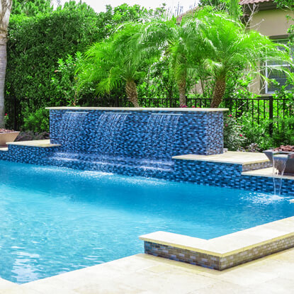 A rectangular backyard pool with a blue mosaic tile water wall feature surrounded by palm trees.