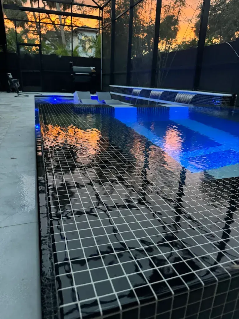 A dark-tiled rectangular pool with a raised spa and water blades under a screened enclosure at dusk.
