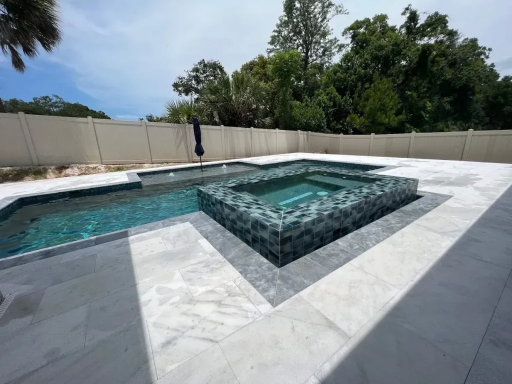A raised dark tiled spa sits beside a pool on a travertine paver patio surrounded by a privacy fence and trees.
