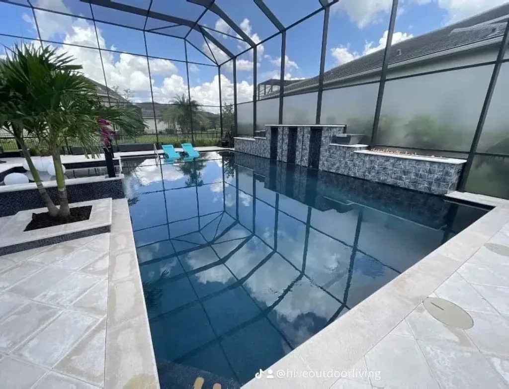 A dark-bottomed rectangular pool under a screen enclosure with a raised stone wall featuring water spouts and fire bowls.